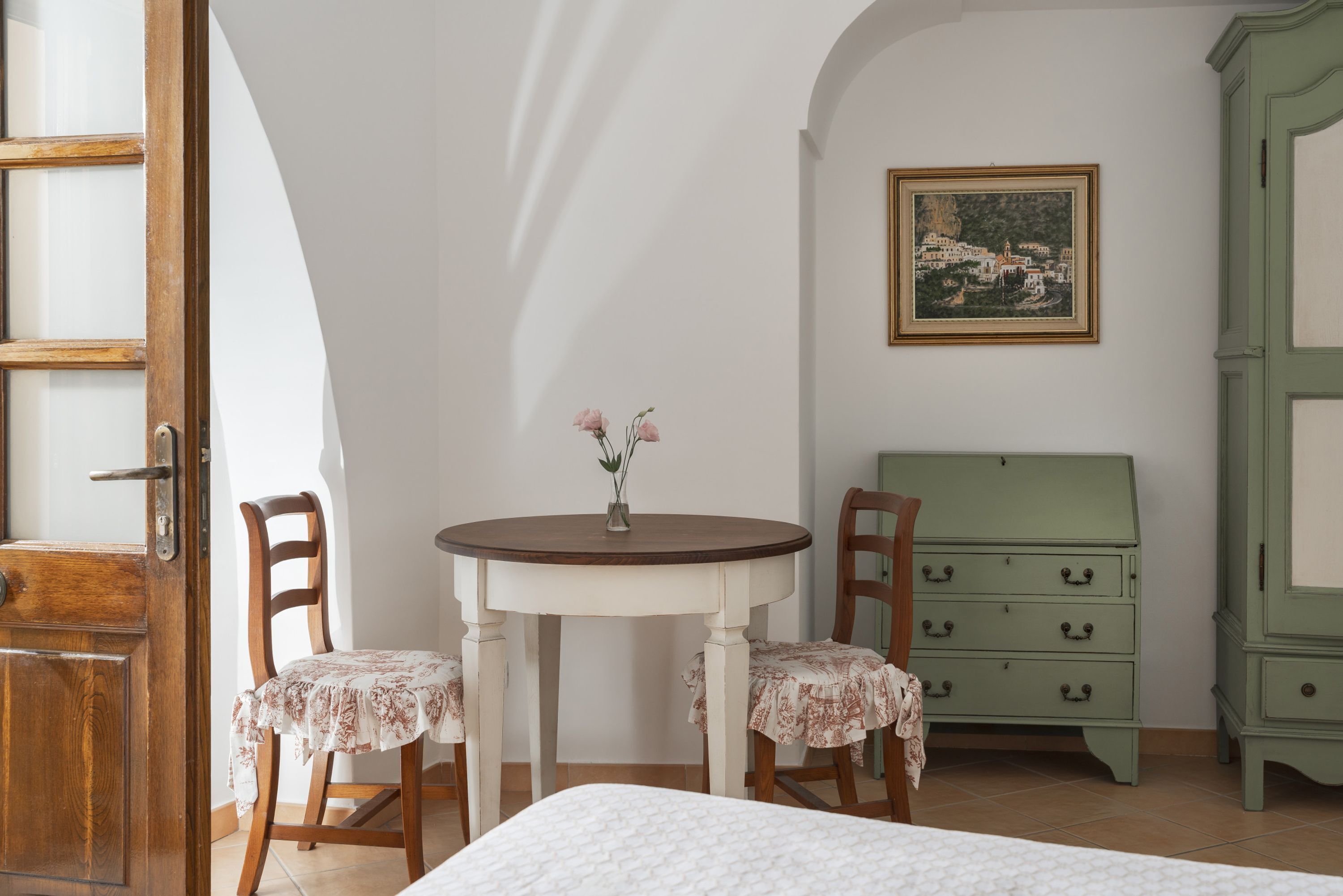 A room with a bed, a wooden chair, a small table with a vase, and a green dresser.