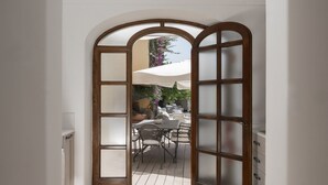 Une porte ouverte donne sur un patio extérieur agrémenté d’une table et de chaises, le tout étant entouré d’un mur blanc et d’un cadre de porte en bois.