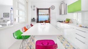 A modern kitchen with a large island, white cabinets, and a view of the sea through an open door. - Villa Mapi in Massa Lubrense (Massa Lubrense)