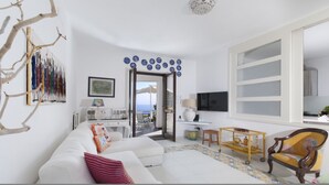 A modern living room with a white sofa, a wooden chair, a small table, and a TV. There is a balcony with a view of the sea. - Villa Mapi in Massa Lubrense (Massa Lubrense)