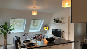 Et moderne køkken med en sort bordplade, to hængende lamper, et spiseområde med en sofa og et stort vindue.