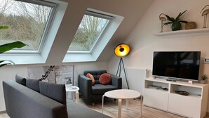 En moderne stue med sofa, lænestol, rundt bord, gulvlampe, fjernsyn og bogreol. Der er ovenlys og udsigt til træer.