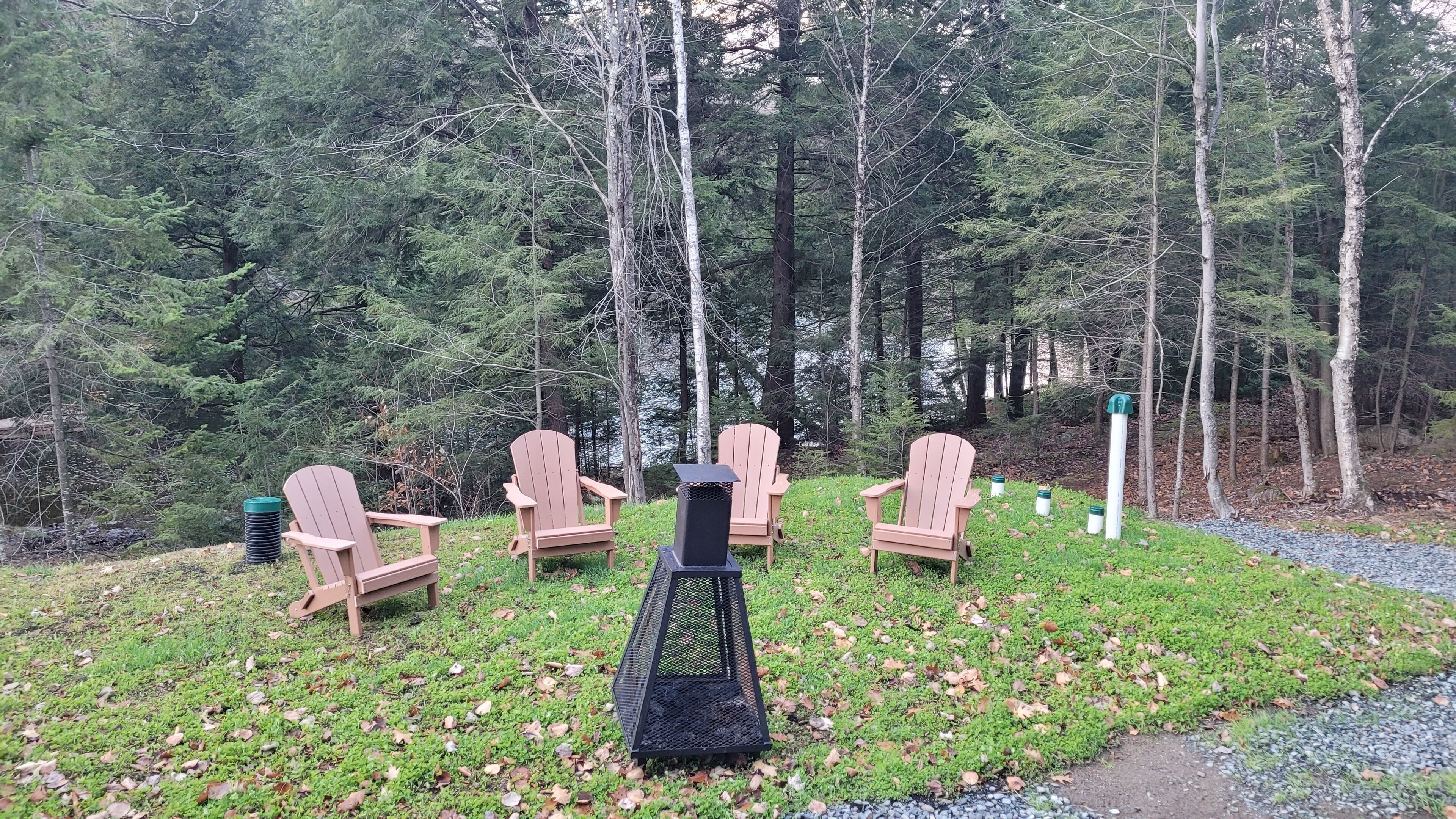 Un grupo de sillas Adirondack de madera dispuestas alrededor de un hogar para fuego en un área boscosa.