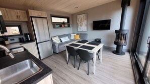 Une cuisine moderne dotée d’un réfrigérateur encastré, un coin repas avec une table et des chaises, un canapé gris agrémenté de coussins à motifs et un poêle à bois noir.