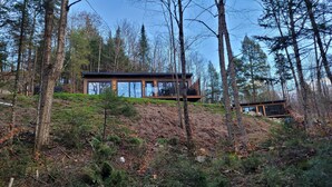 Une cabane moderne dotée de grandes fenêtres, entourée d’une forêt dense et située au pied d’une colline.