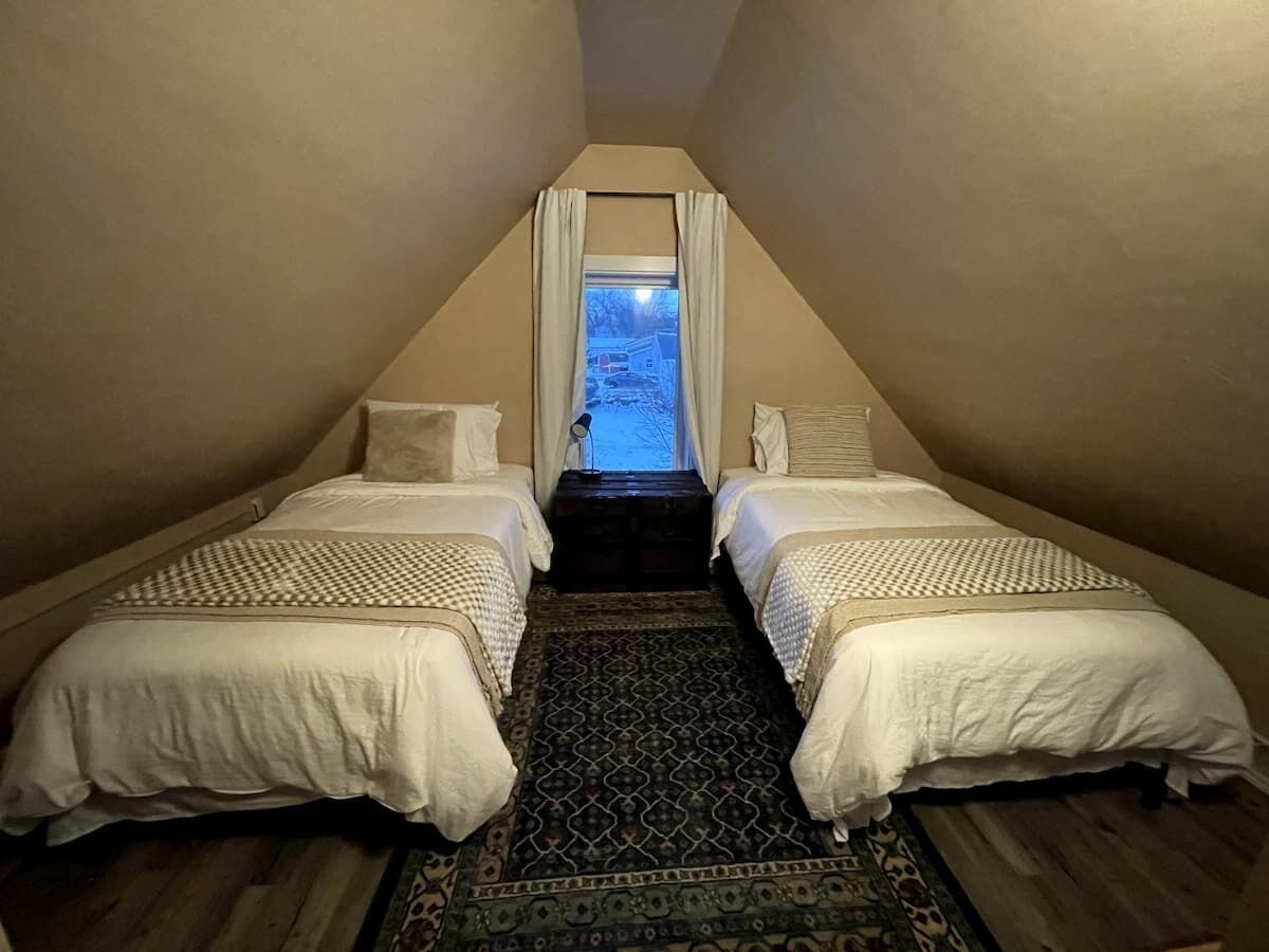 A narrow attic bedroom with two beds, a patterned rug, and a view through a window.