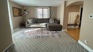 Una sala de estar con sofá, reposapiés y alfombra estampada. Hay una ventana con persianas, una lámpara y un perchero.