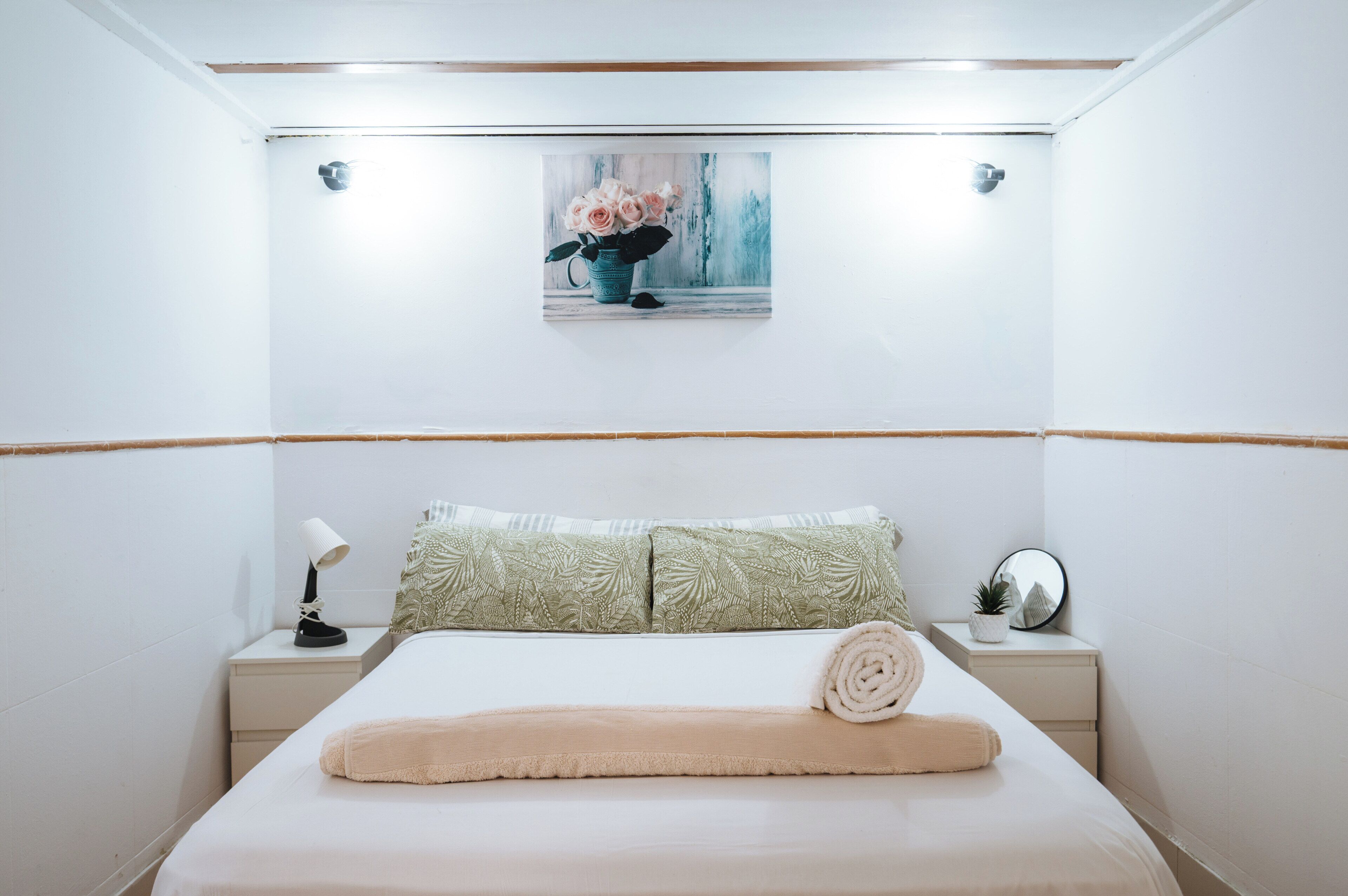 A neatly arranged bedroom with a bed, pillows, a bedside lamp, a small table with a mirror, and a wall-mounted picture.