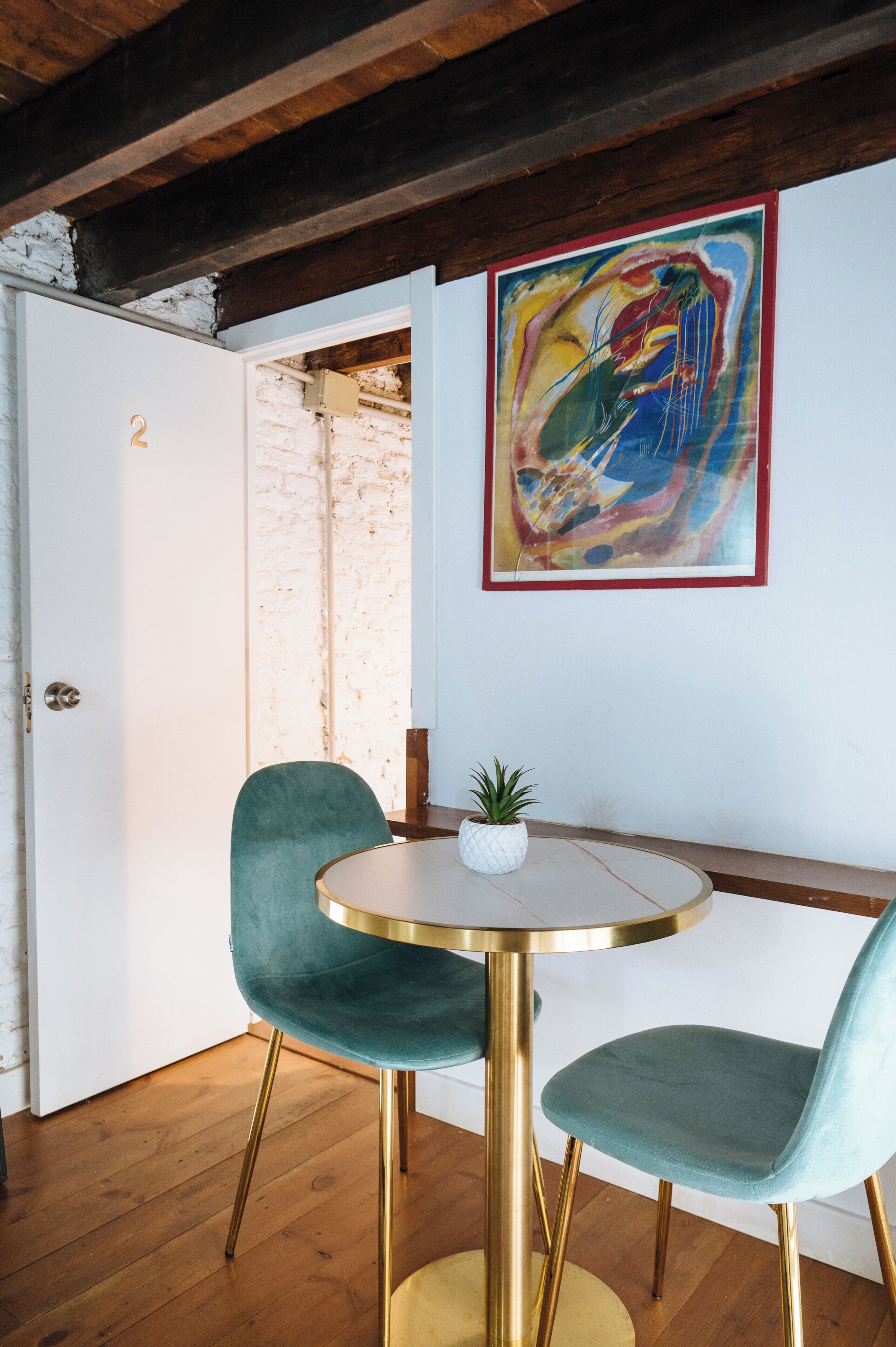 A room with a round table, two teal chairs, a single potted plant, a colorful abstract painting, and exposed brick walls.