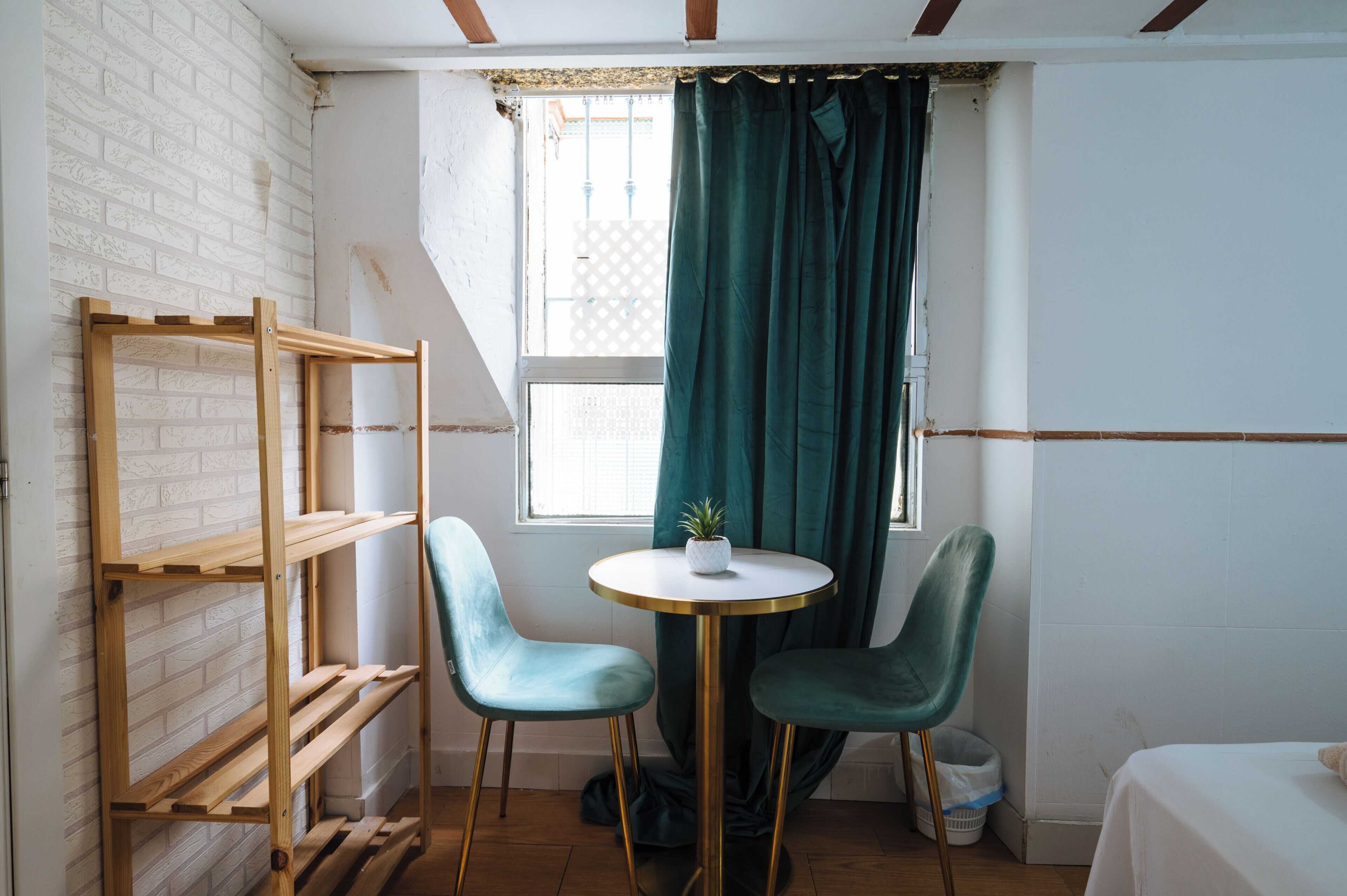 Una habitación con pared de ladrillo, ventana con cortinas, una pequeña mesa redonda y dos sillas, y una cama.