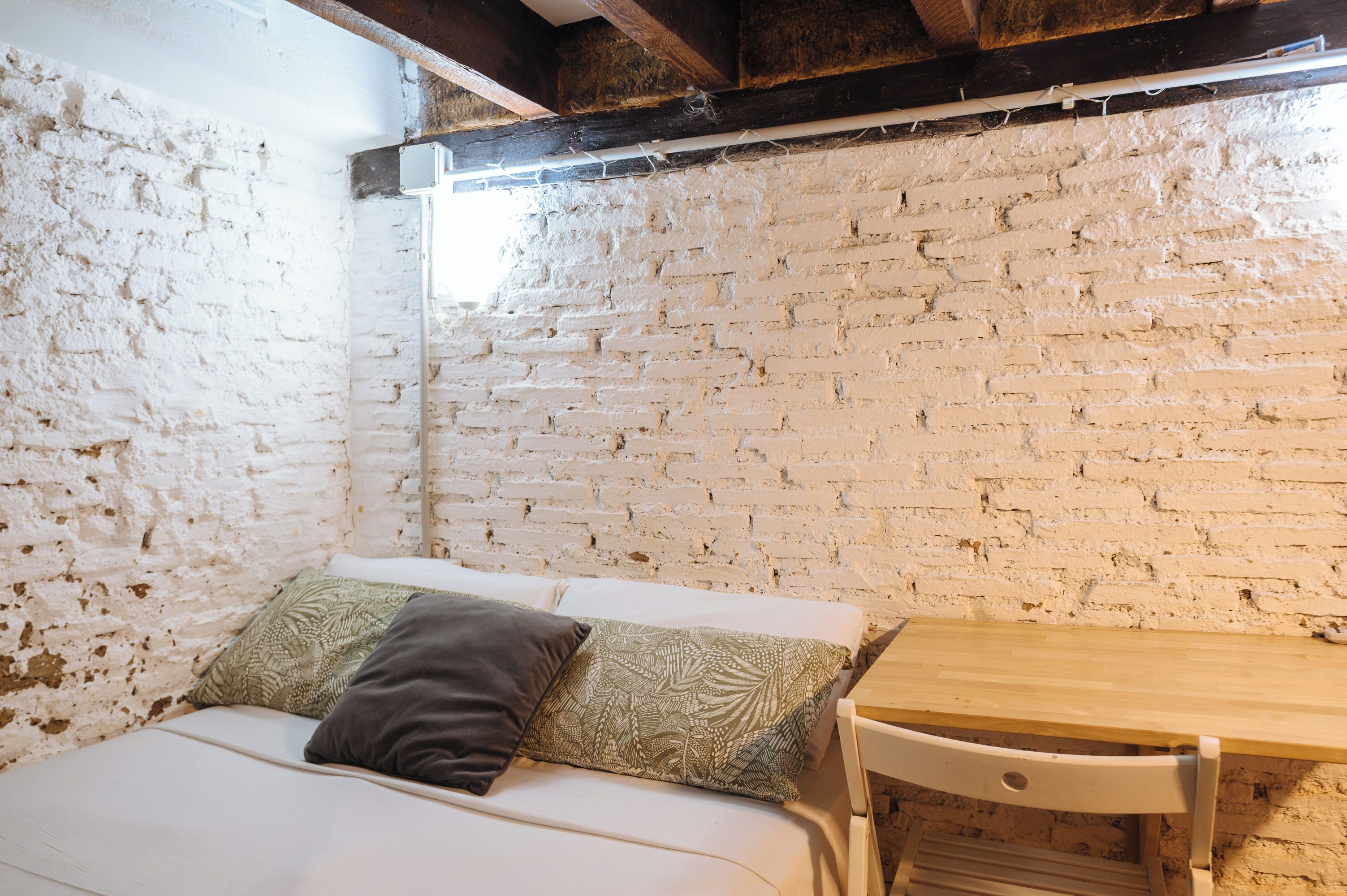 Una habitación con pared de ladrillo blanco, viga de techo de madera, cama blanca con almohadones estampados y una mesa de madera.
