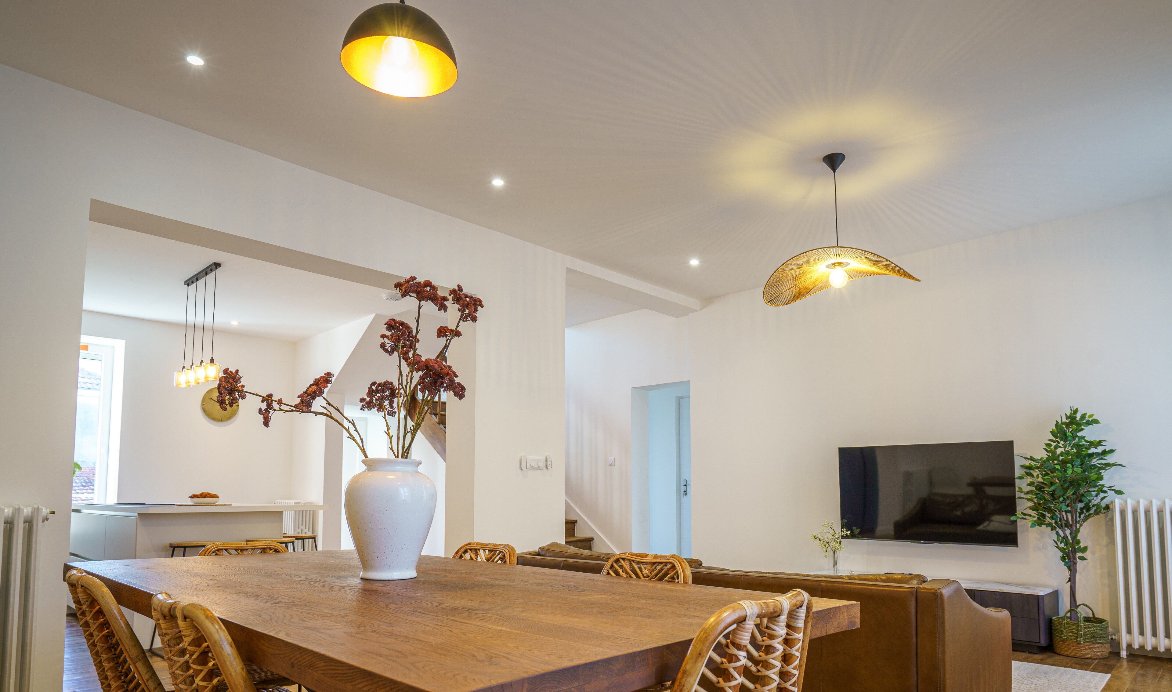 Uma sala de jantar moderna com mesa de madeira, cadeiras de vime, um vaso branco com flores e iluminação pendente.
