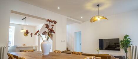 Uma sala de jantar moderna com mesa de madeira, cadeiras de vime, um vaso branco com flores e iluminação pendente.