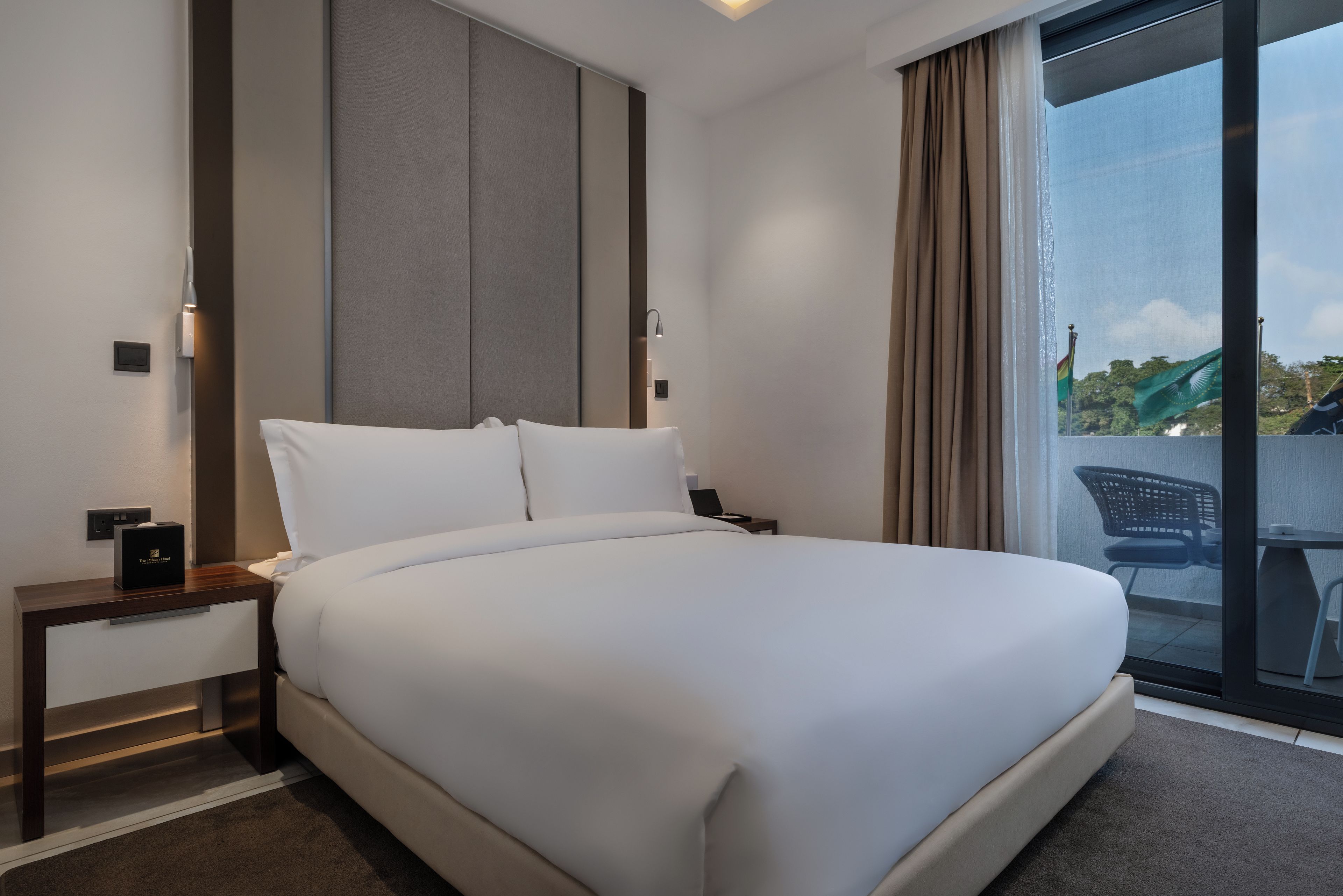 A hotel room with a large bed, a bedside table, a nightstand, a window with curtains, and a view of the outside.