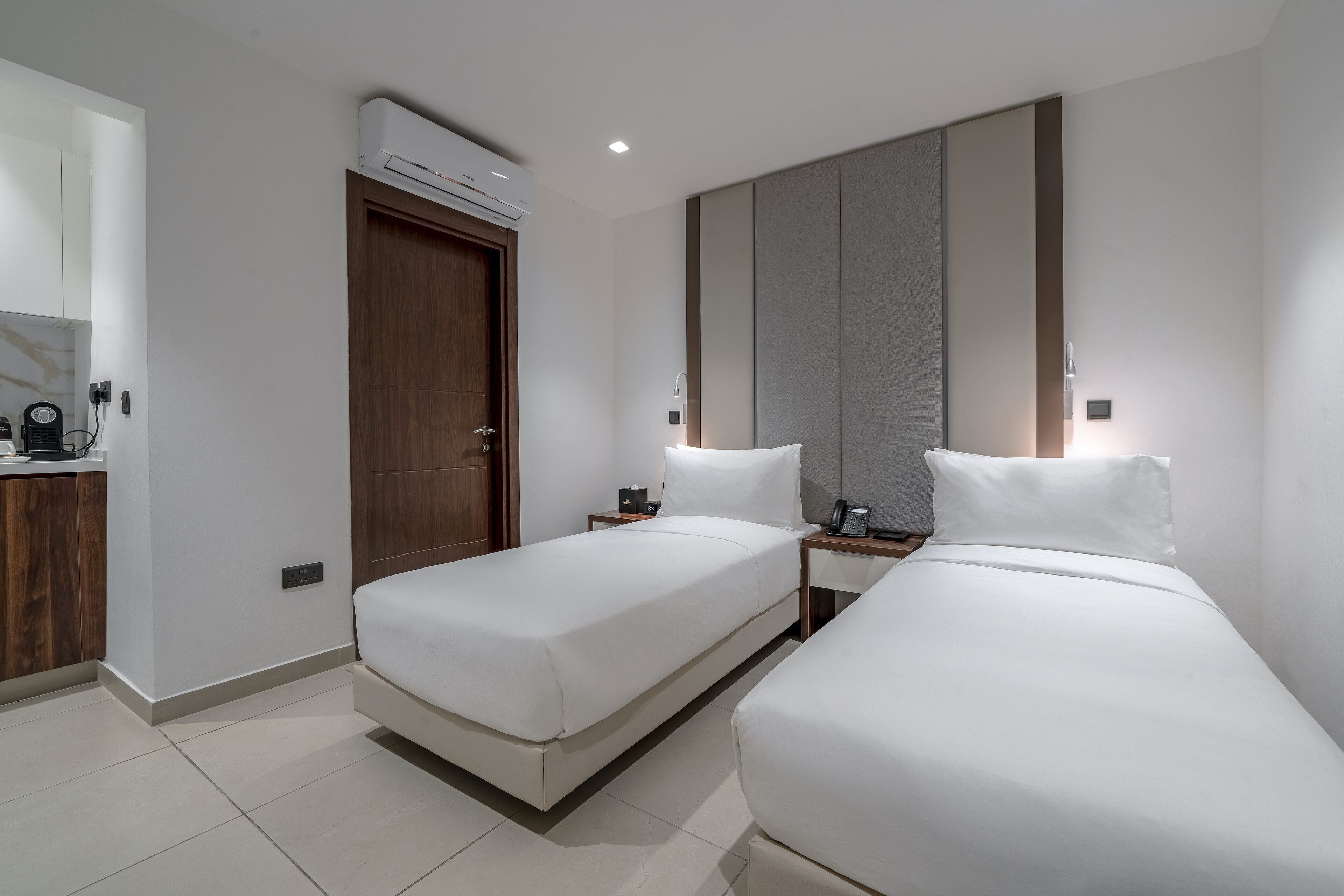 A hotel room with two single beds, a wooden wardrobe, a mirror, and a small table with a phone and a cup.