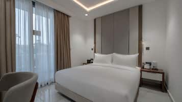 A modern hotel room with a large bed, a bedside table with a phone and lamp, a chair, and a view through sheer curtains.