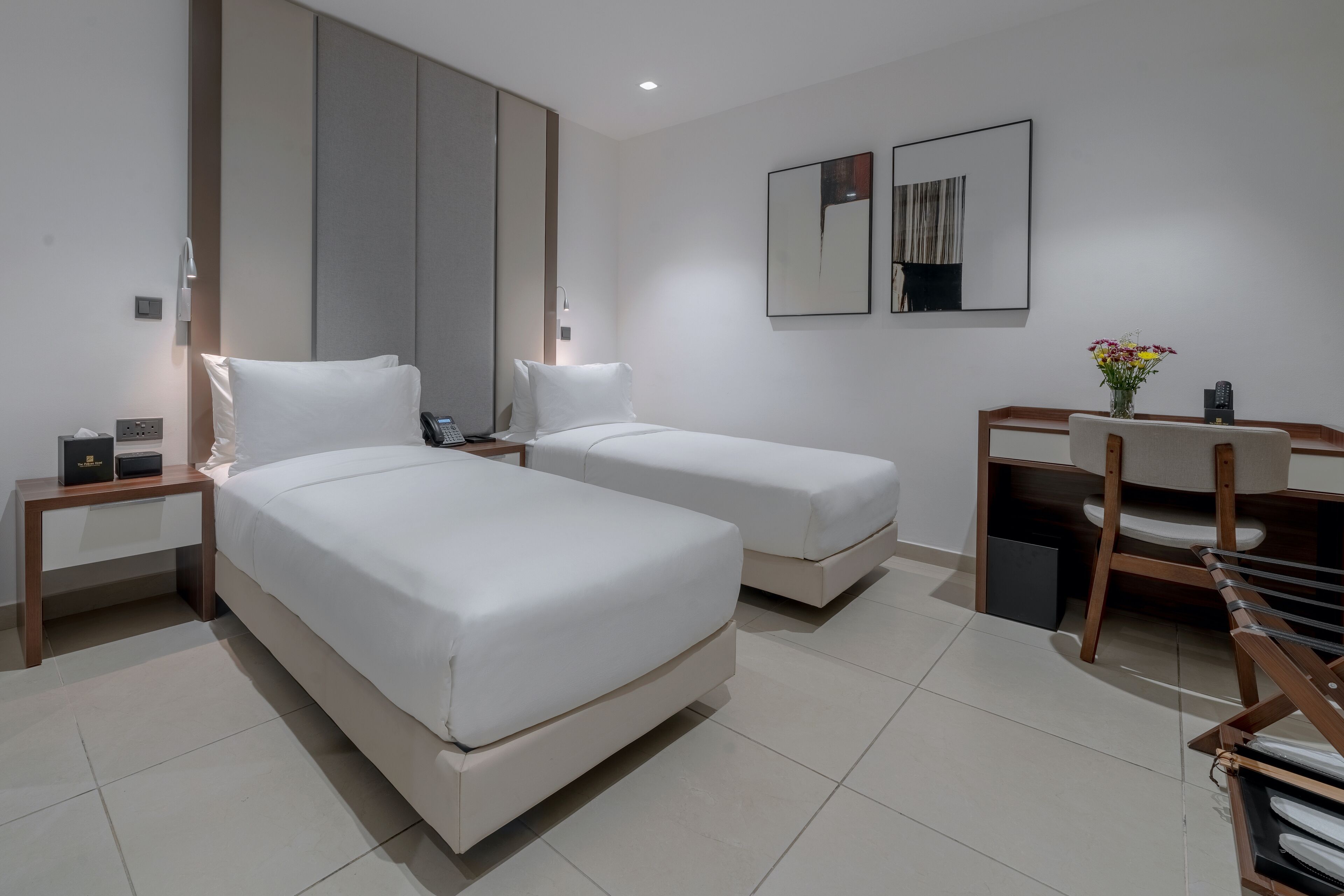 A modern hotel room with two single beds, a desk, a chair, and a small table with a vase of flowers.