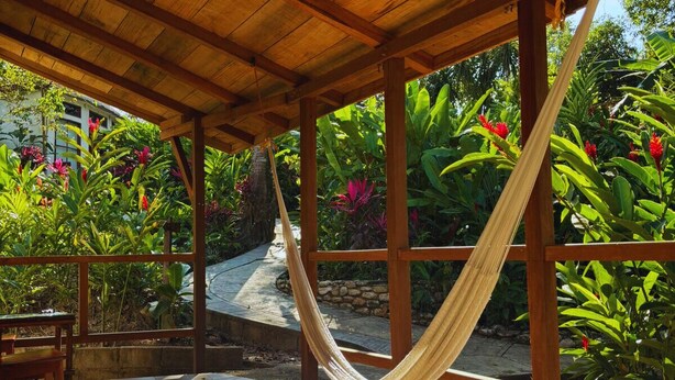 Uma pérgola de madeira com uma rede, rodeada por plantas tropicais exuberantes e um pátio com ladrilhos.