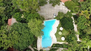 Vista aérea de uma propriedade com piscina, rodeada por vegetação exuberante, área pavimentada com um carro e um edifício com telhado vermelho.