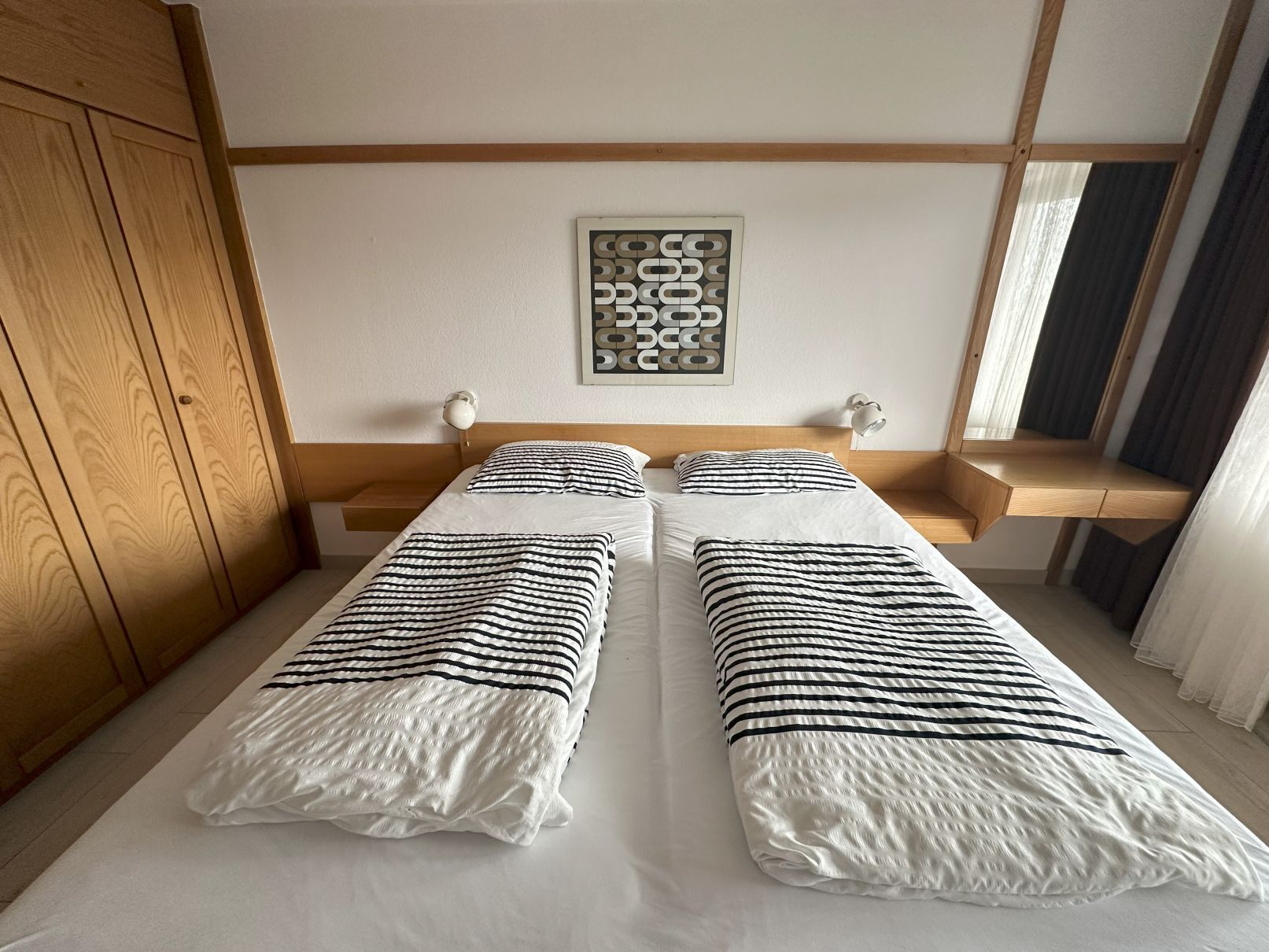 Una cama doble con ropa de cama a rayas, un cabecero de madera, una obra de arte abstracto colgada en la pared y una mesita de noche con una lámpara.