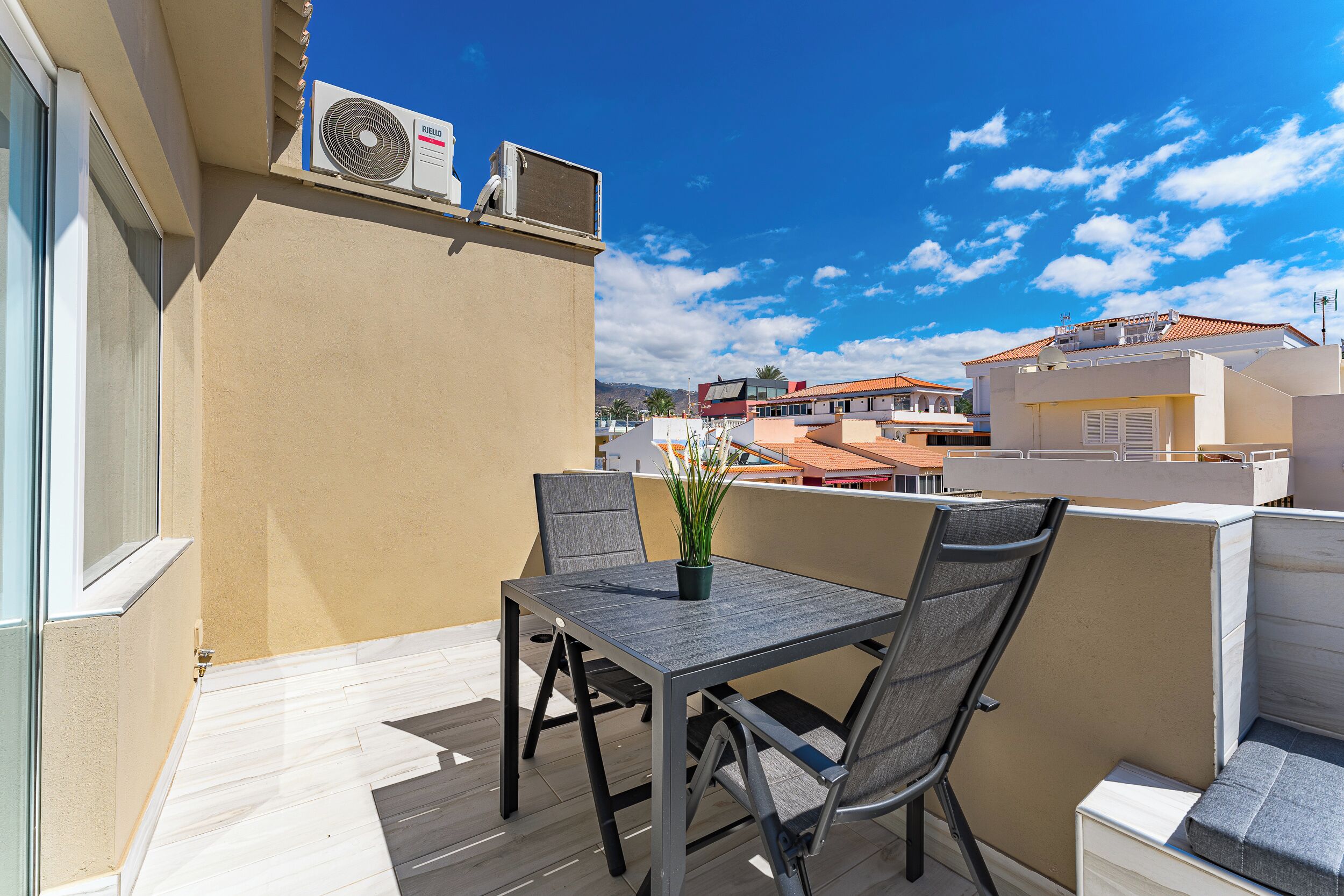 Un patio al aire libre con una mesa y sillas, una planta en maceta y un equipo de aire acondicionado en la pared.