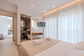 A modern hotel room with a large bed, a bathroom with a marble wall, a mounted TV, and a large window with sheer curtains.