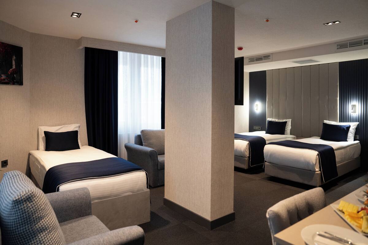 A hotel room with two beds, a sofa, a dining table with a plate of fruit, and a large window with curtains.