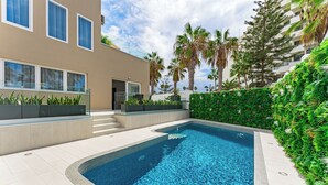 A modern pool area with a clear blue pool, surrounded by a low hedge and a building with large windows. - Bal One bedroom apartment Adults Only (204) (Arona)