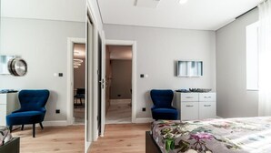 A modern bedroom with a wooden floor, a bed with a floral bedspread, two blue chairs, a white dresser, and a small desk with a mirror.