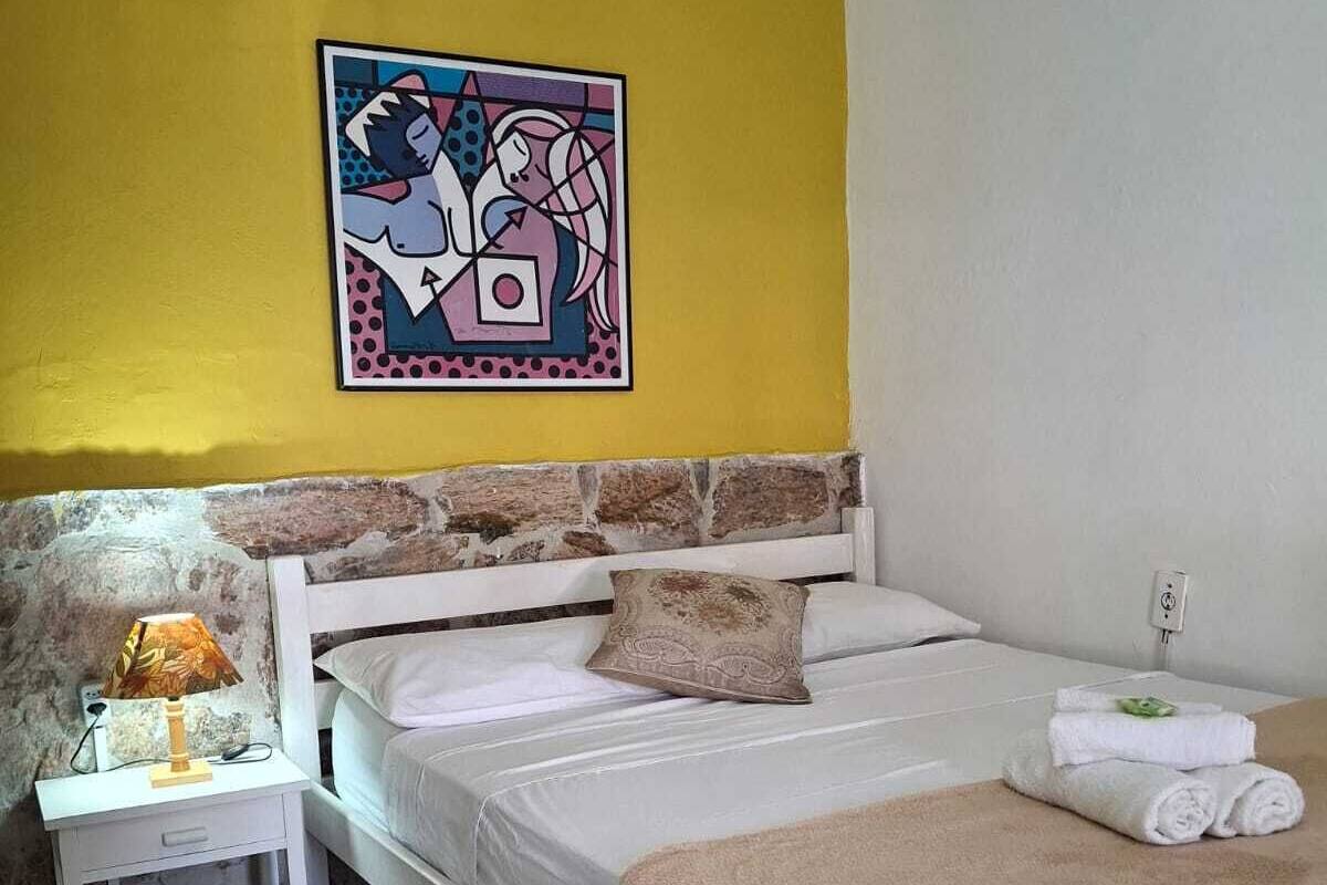 A bedroom with a bed, a nightstand, a lamp, a painting, and a tiled floor.