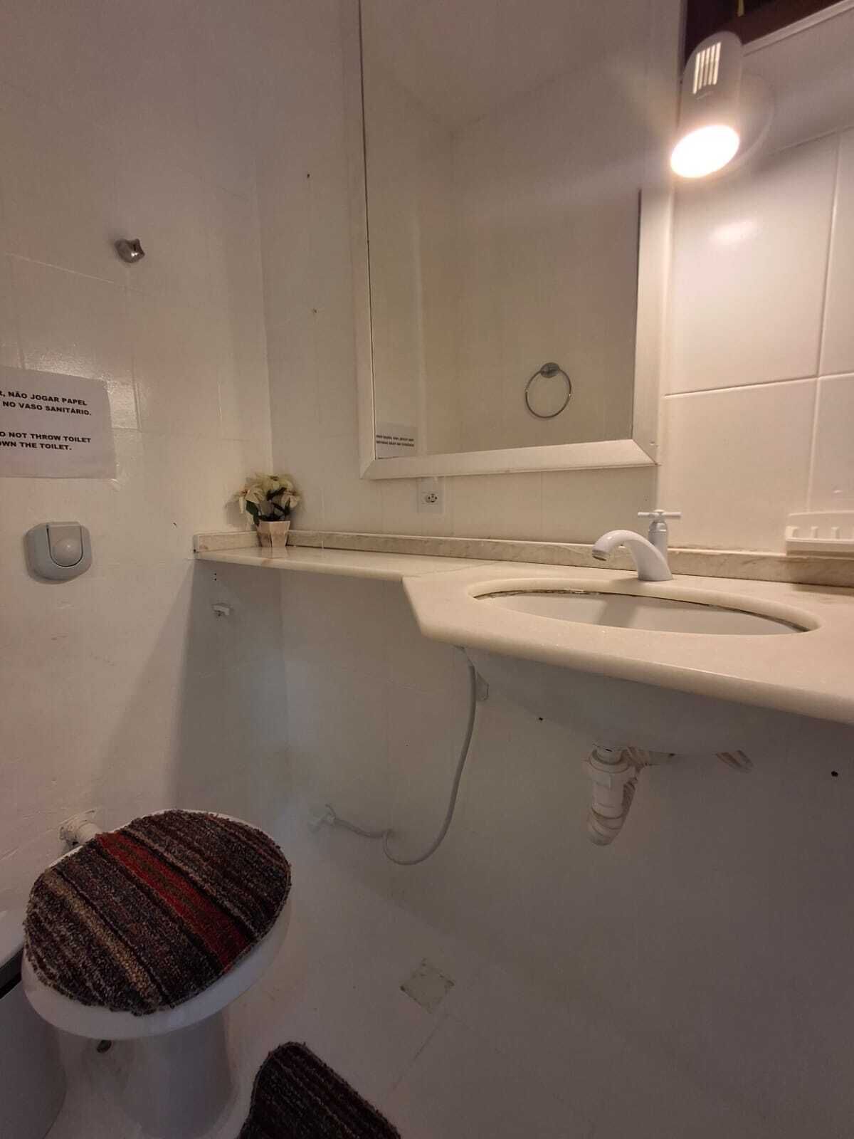 A bathroom with a toilet, sink, and mirror. There is a sign on the wall and a small flower arrangement on the sink counter.