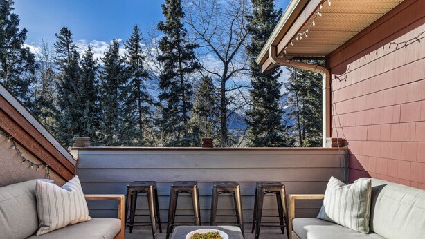 Un espace extérieur aménagé avec deux canapés et quatre tabourets, une table ornée d’un vase rempli de verdure, et une vue sur de grands arbres.