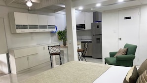 A modern hotel room with a bed, a green armchair, a dining area with a table and chairs, and a kitchenette.