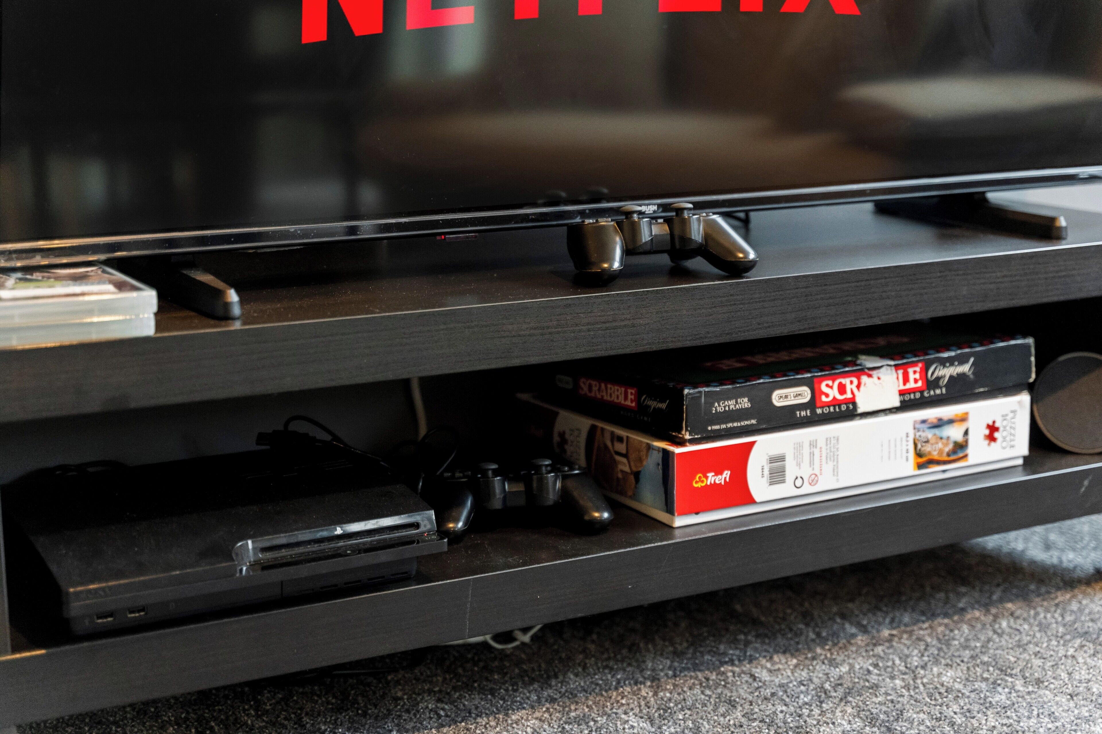 A television on a stand with a remote control, a game controller, and a Scrabble box underneath.