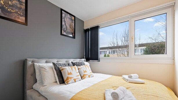 A neatly made bed with a yellow blanket and white towels, a grey wall with a framed picture, and a window with curtains. - Charming 3 Bedroom House in Birmingham Sleeps 7 (Birmingham)