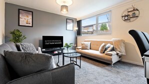 A modern living room with a grey sofa, a black armchair, a glass coffee table, a flat-screen TV, and a large window offering a view of residential buildings. - Charming 3 Bedroom House in Birmingham Sleeps 7 (Birmingham)