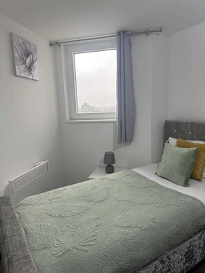 A bedroom with a bed, a window with curtains, a radiator, a bedside lamp, and a wall-mounted artwork. - 1br-sleeps4-parking-closetocentre (Birmingham)
