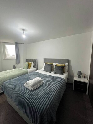A double bed with a grey headboard, two bedside tables, a window with curtains, and a bedside lamp. - 1br-sleeps4-parking-closetocentre (Birmingham)