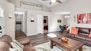 A spacious living room with a brown leather sofa, a wooden coffee table, and a ceiling fan. A kitchen is visible in the background, and a dining area with a table and chairs is also present.