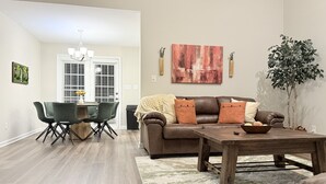 A living room with a brown leather sofa, a wooden coffee table, and a dining area with green chairs and a wooden table.