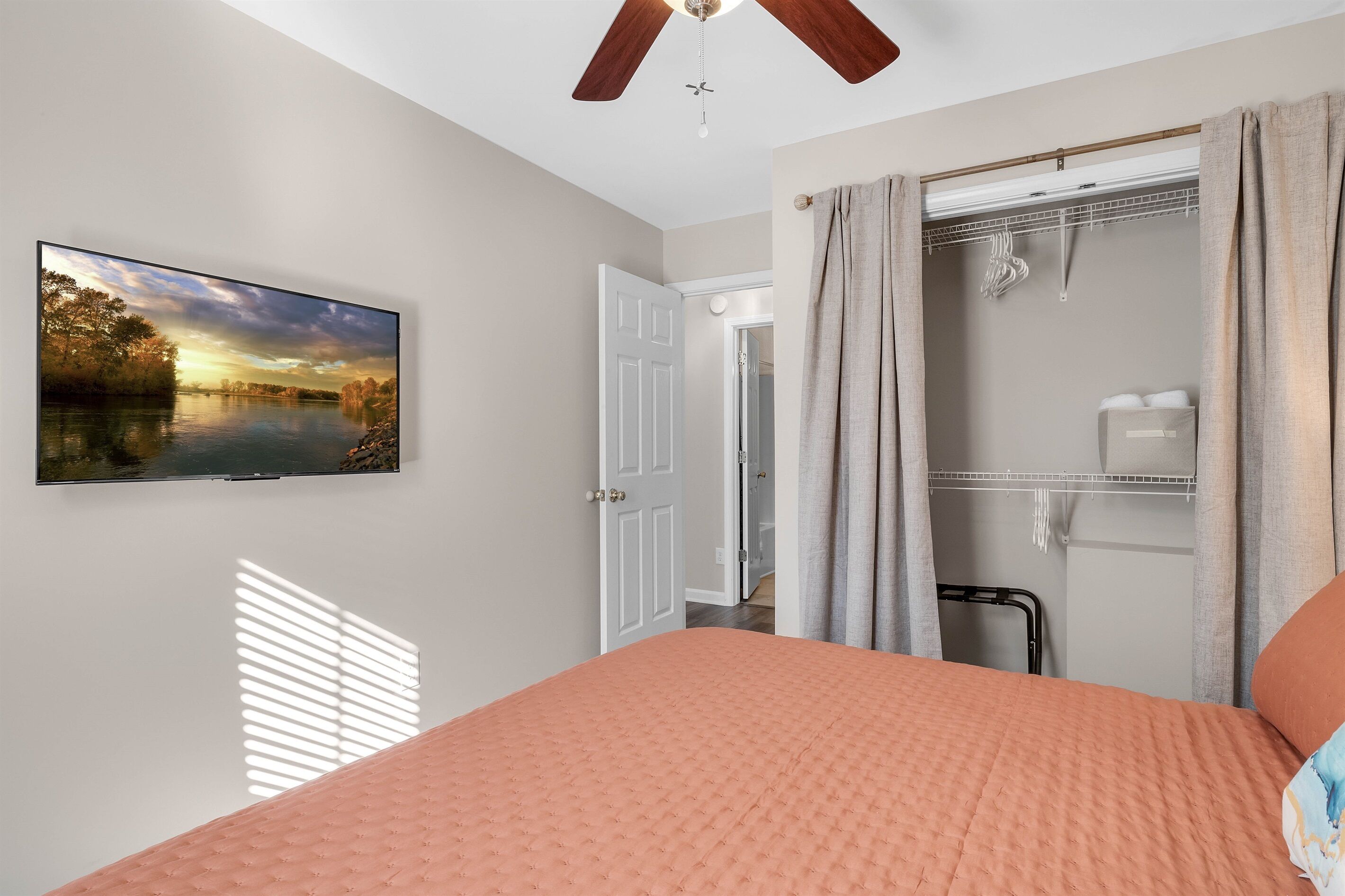 A bedroom with a bed, a TV mounted on the wall, a ceiling fan, and a walk-in closet with a mirror and hanging clothes.