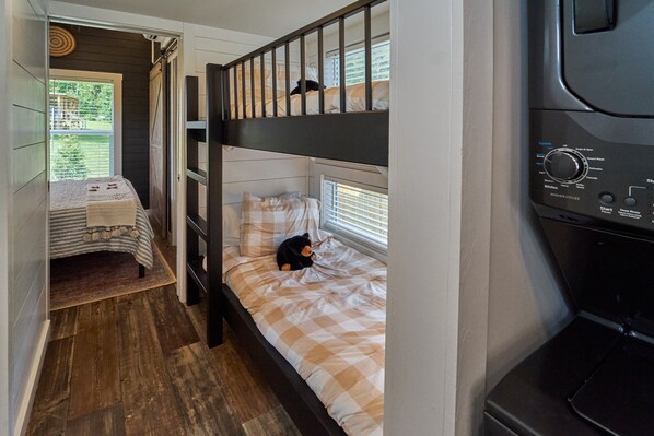 A compact room with a bunk bed, a small table with a tablecloth, and a washing machine. - Lil Lake House! Modern Tiny Home! (Sevierville)