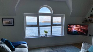 Ein Wohnzimmer mit einem großen Bogenfenster mit Meerblick, einem am Wand montierten Flachbildfernseher und einer gemütlichen Sitzecke mit einem grauen Sofa und gemusterten Kissen.