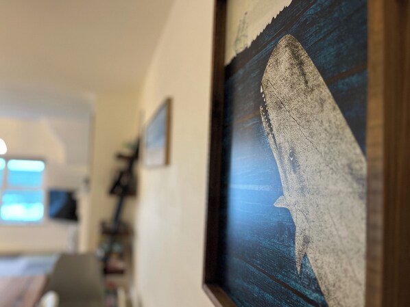 Une œuvre d’art encadrée représentant un requin, accrochée à un mur dans une pièce dotée d’une fenêtre et d’une porte en arrière-plan.