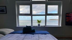 Ein Schlafzimmer mit einem Bett, einem Fenster mit Meerblick, einer Topfpflanze und einem gerahmten Bild an der Wand.
