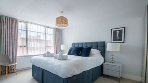 A modern bedroom with a large bed, a bedside table, a chair, and a window with curtains. - Cockleshells - Modern apartment in Swanage (Swanage)