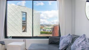 A modern living room with a grey sofa, a white ottoman, and a large window offering a view of a building and a parking area. - Cockleshells - Modern apartment in Swanage (Swanage)