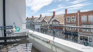 A balcony with a table and chairs, overlooking a street with buildings and shops. - Cockleshells - Modern apartment in Swanage (Swanage)