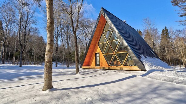 A modern, triangular cabin with large windows, surrounded by a snowy forest.
