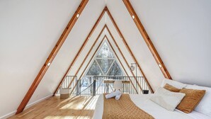 A bedroom with a sloped ceiling, wooden beams, a bed with white bedding, and a view of trees through a large window.